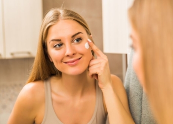 Jovem mulher na frente do espelho aplicando hidratante facial na pele oleosa.
